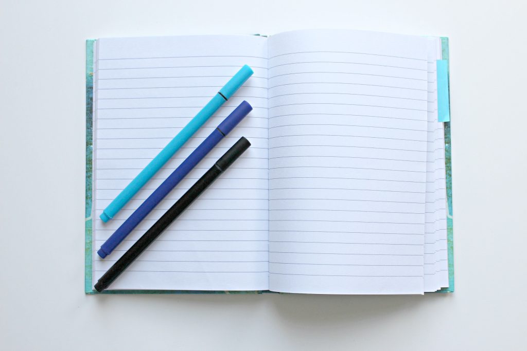 Open lined notebook with colorful pens on a white surface, perfect for note-taking and office tasks.